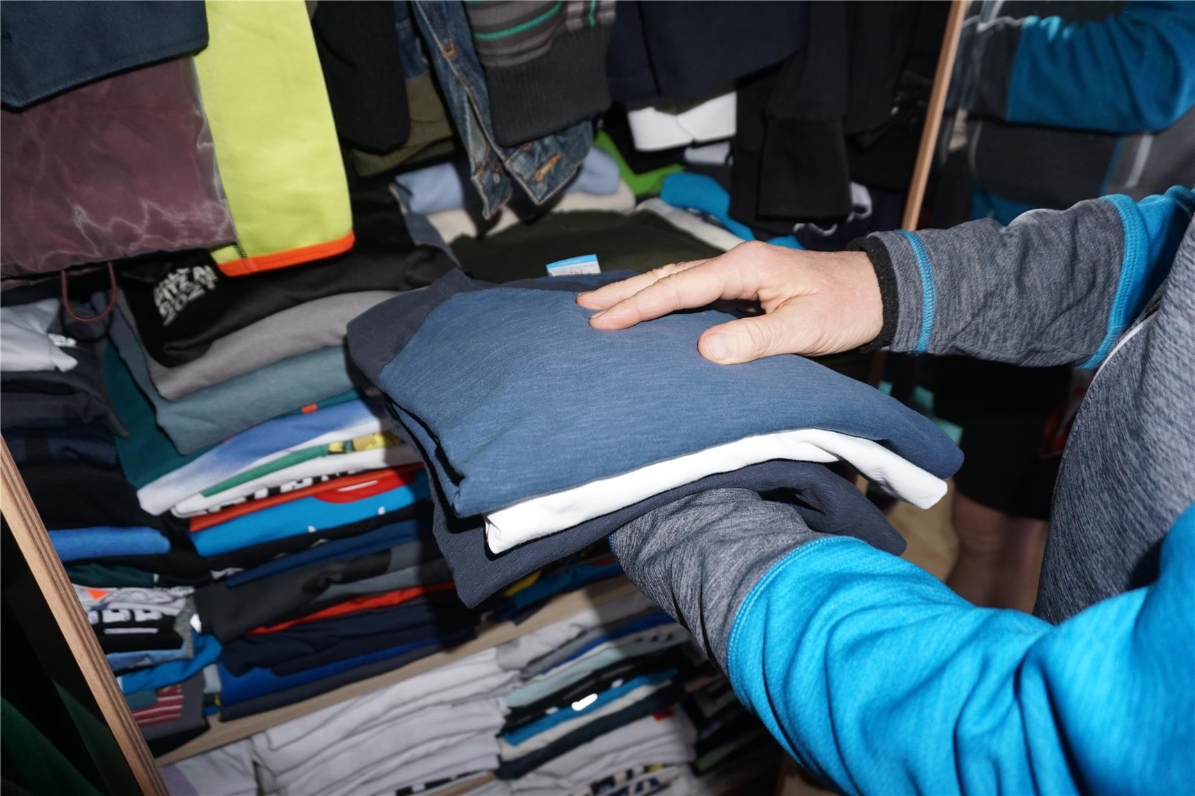 Zwei Hände halten einen Stapel T-Shirts vor einem geöffneten Kleiderschrank.