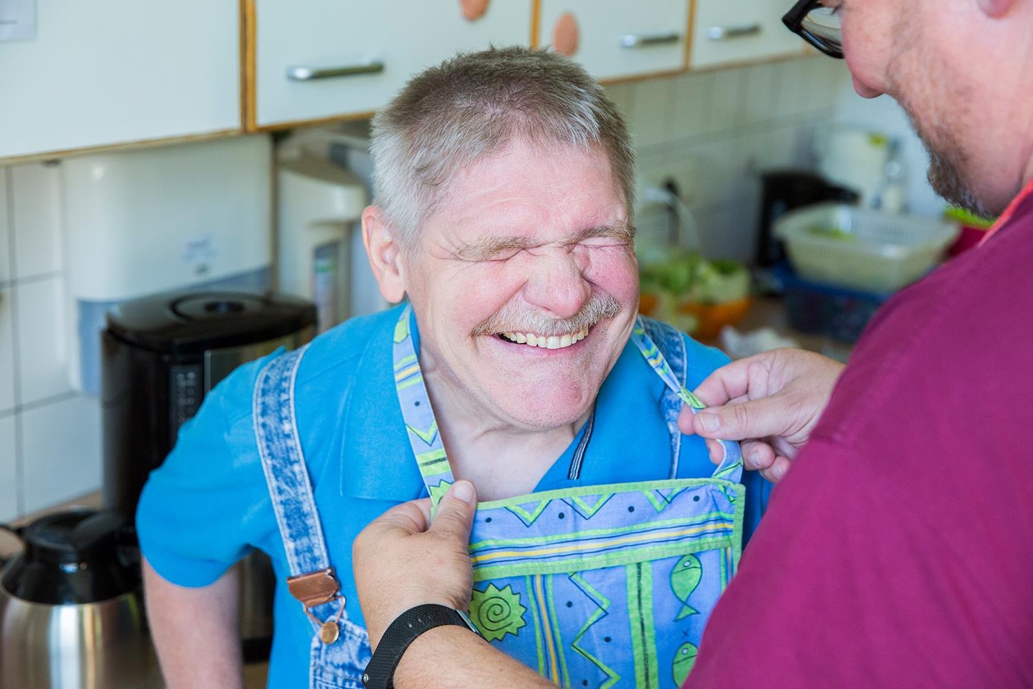 Ein Mann mit Behinderung lacht während einen Latz angelegt bekommt (Deutscher Caritasverband e. V. / Sebastian Pfütze)