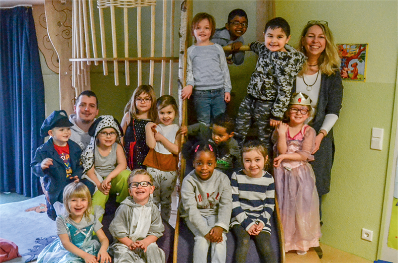 Gruppenfoto mit einer Betreuerin, einem Betreuer und 13 Kindern in einem Spielzimmer einer Kindertageseinrichtung