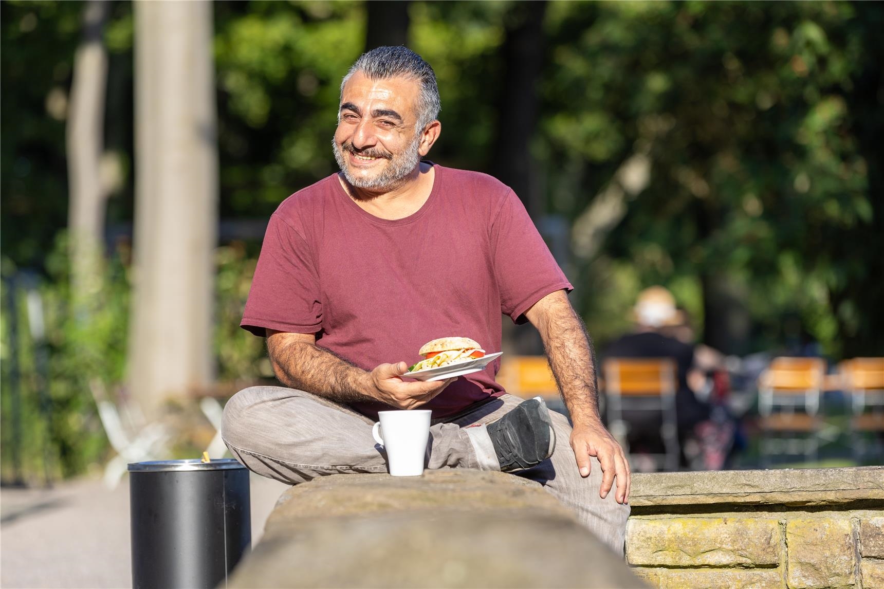 Ein Mann sitzt auf einer Mauer mit einem Brötchen und einer Kaffeetasse. 