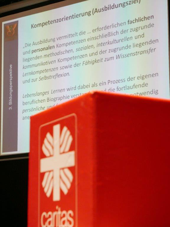 Bühne mit Leinwand auf der Informationsveranstaltung in Dortmund. Auf der Leinwand wird eine Präsentation gezeigt, im Vordergrund ist ein roter Caritas-Würfel zu sehen. (Jürgen Sauer)