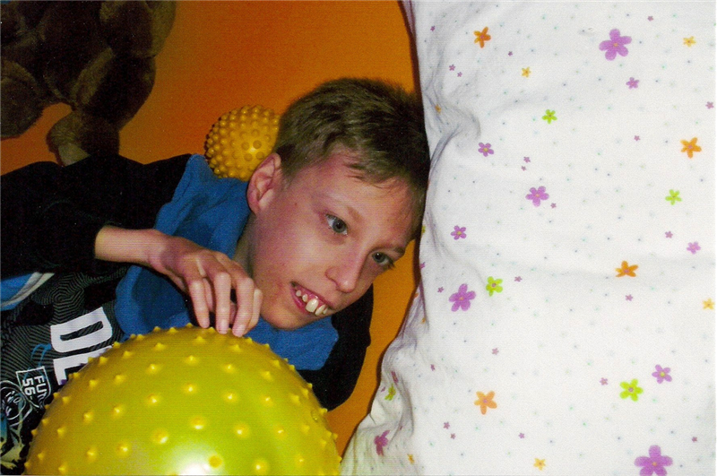 Ein Junge mit Behinderung, der in einem Snoezelraum liegt. Um ihn herum liegen zwei Massagebälle, ein Kuscheltier und eine Decke. (Caritas)