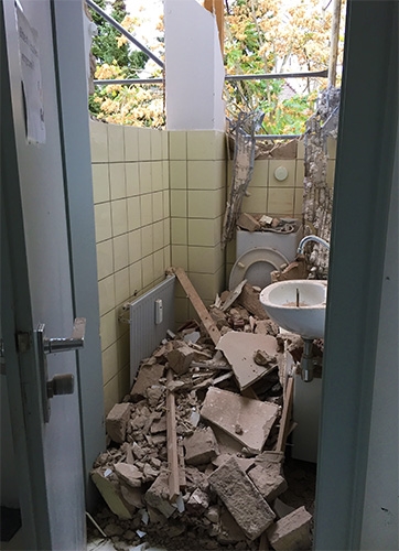 Gefliester Toilettenraum, teilweise abgerissen, Bauschutt am Boden, durch die teilweise fehlende Wand sind außen die Bäume sichtbar (Caritasverband Darmstadt e. V. / M. K. Triebel)