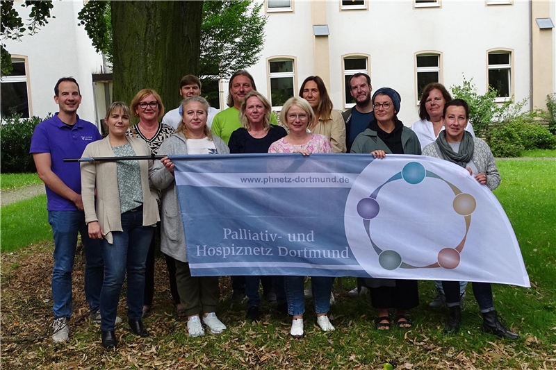 Die Mitglieder des Palliativ- und Hospiznetz Dortmund im Gruppenbild. Sie halten eine Beachflag mit dem Logo in den Händen.