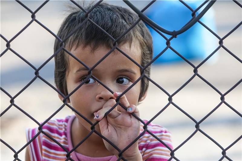 Ein Flüchtlingskind schaut durch einen Maschendrahtzaun. 
