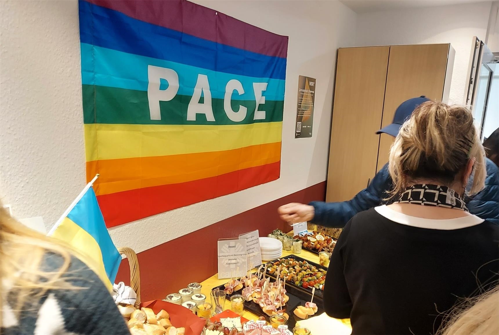 Regenbogenflagge mit Schriftzug Frieden "Pace" vor einem Buffet mit Personen 