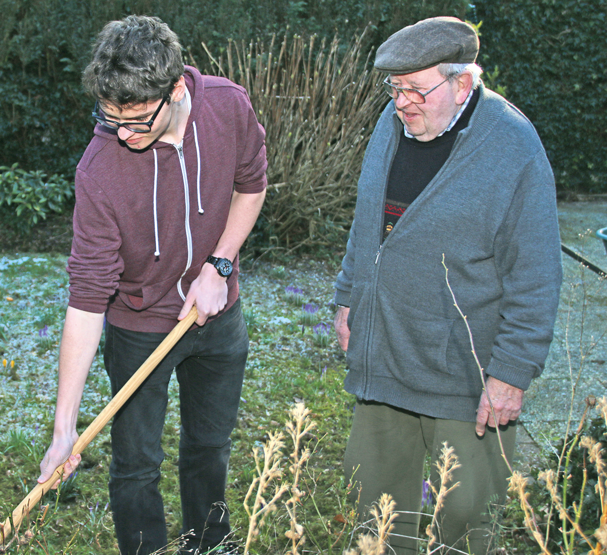 Ein junger Mann bearbeitet mit einem Arbeitsgerät die Wiese eines Gartens. Ein älterer Mann steht dahinter und schaut zu. (Julius Schwerdt)
