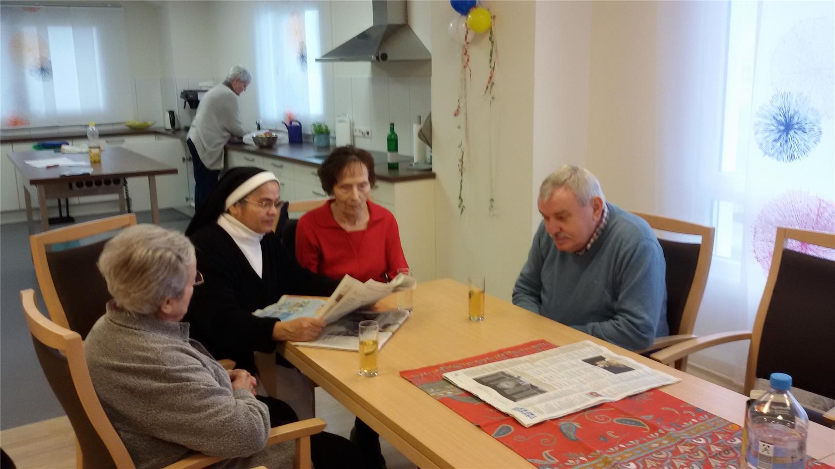 Um einen Tisch in der Tagespflege sitzen zwei Damen und ein Herr, ein Ordensschwester liest vor. Im Hintergrund arbeitet jemand im Küchenbereich. (© Caritasverband Worms e. V.)