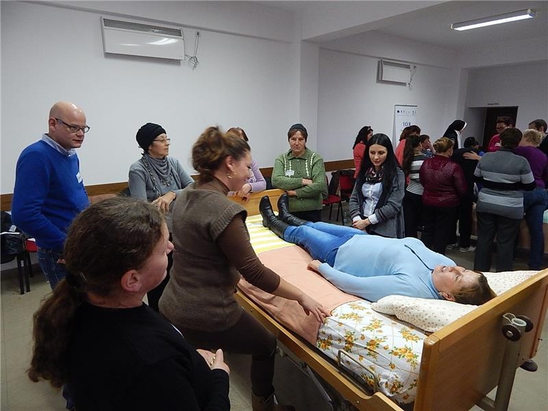 Zentral im Bild ist ein Krankenbett zu sehen, auf dem eine Frau liegt. Um das Bett herum stehen mehrere Personen. (Caritas)