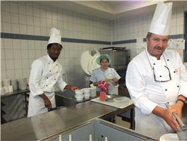 In einer Großküche stehen ein junger, dunkelhäutiger Mann, eine Frau und der Küchenchef mit Kochmütze / Foto: Schedlbauer