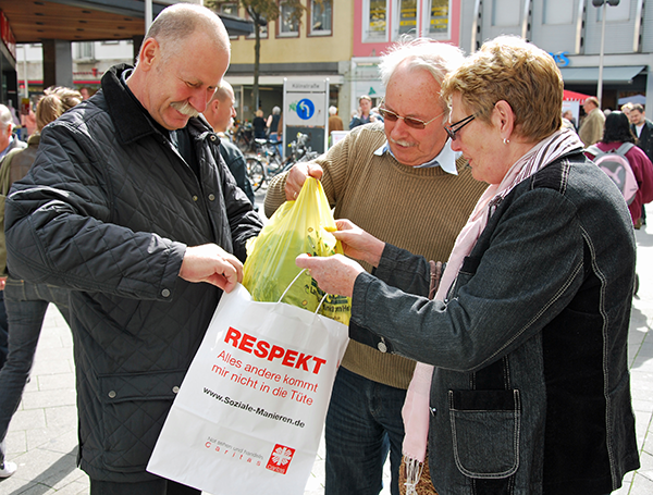 Gerd Schnitzler (links) hilft zwei Passanten in einer Fußgängerzone eine Einkaufstüte in eine Respekttüte umzupacken (Caritas)