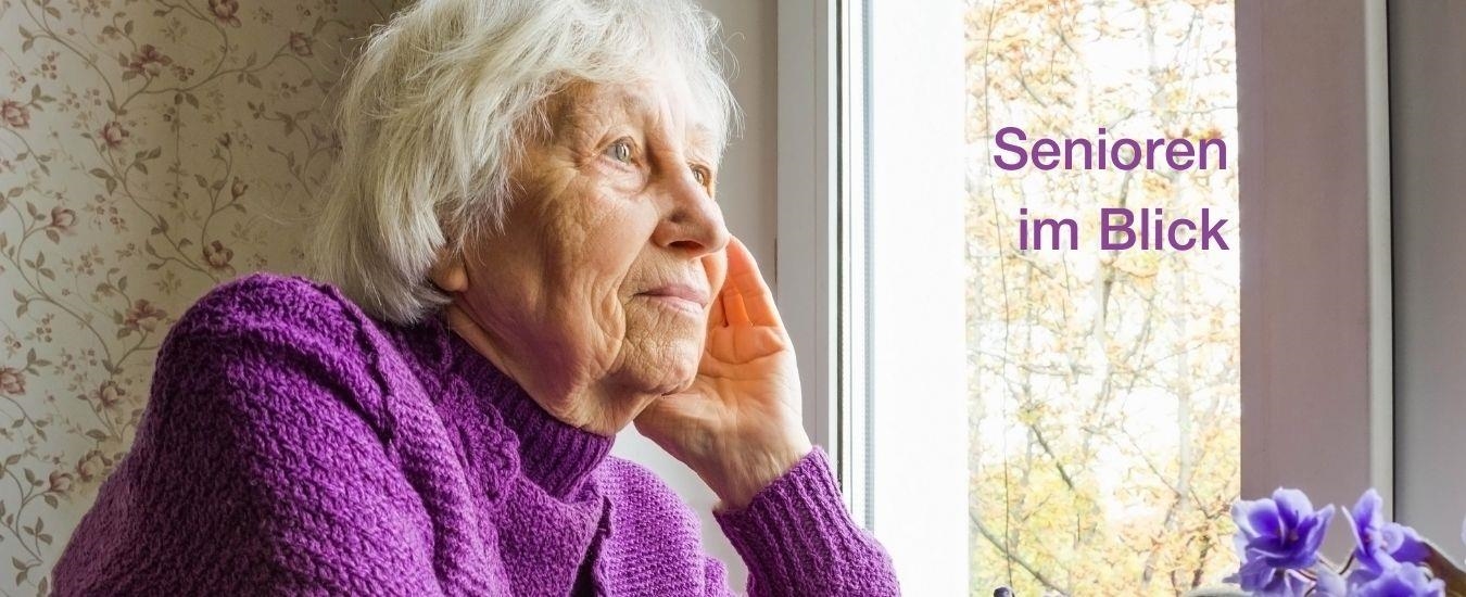 Eine alte Frau mit weißen Haaren und in einem violetten Pullover schaut gedankenverloren aus einem Fenster.