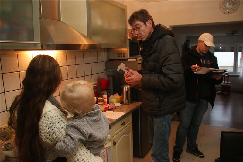 Das Foto zeigt zwei Männer und eine junge Frau mit Baby auf dem Arm in einer Küche.