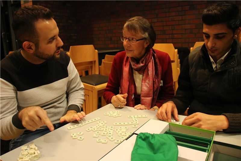 Das Foto zeigt zwei junge Flüchtlinge mit einer Frau, die an einem Tisch Scrabble spielen und sich unterhalten.