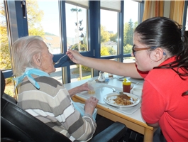 Eine junge Frau sitzt am Tisch mit einer Seniorin und gibt ihr mit einem Löffel Essen ein / Foto: Schedlbauer
