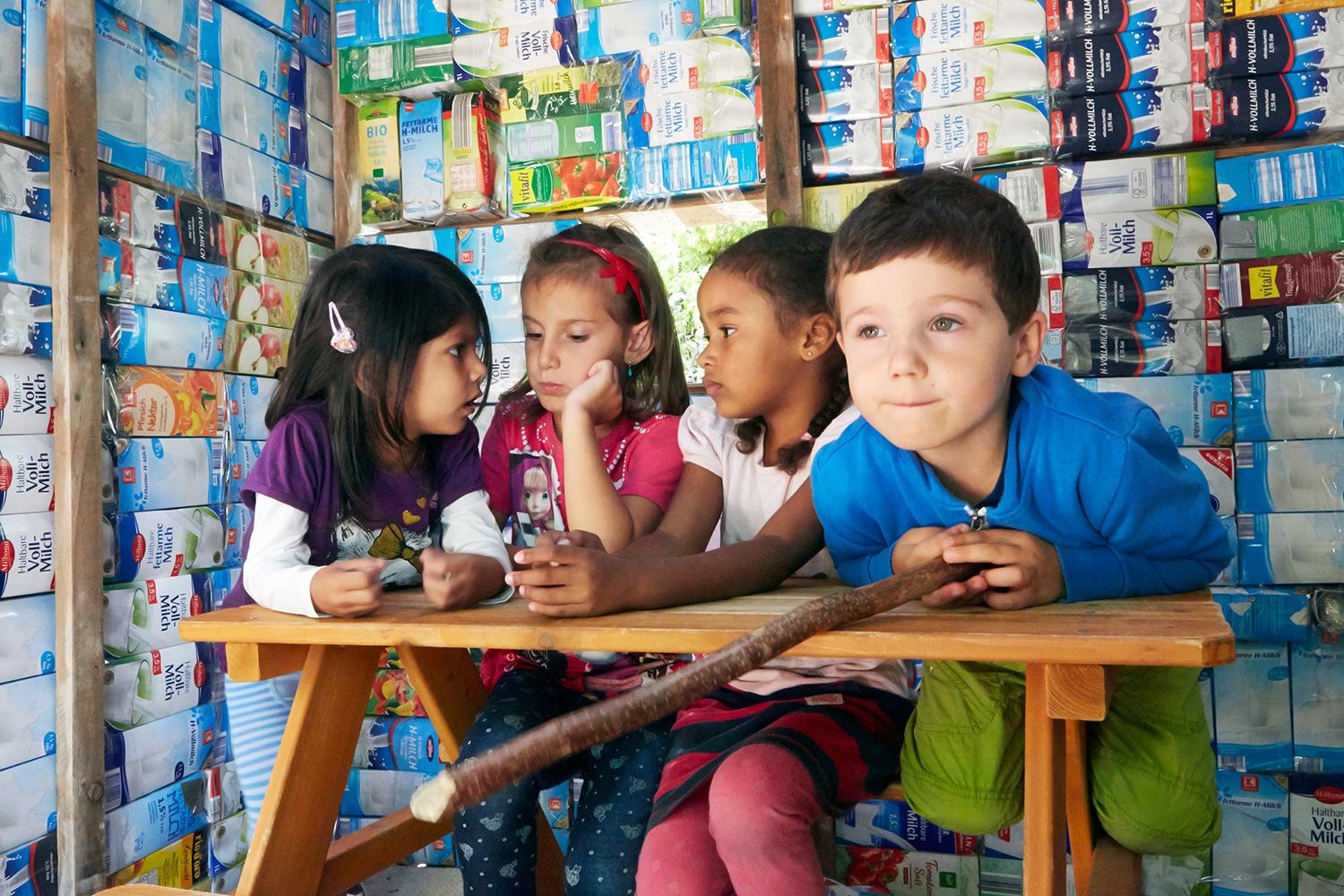 Vier Kinder sitzen an einem Tisch, im Hintergrund eine künstlerische Wand aus Milchtüten. (Birgit Betzelt)