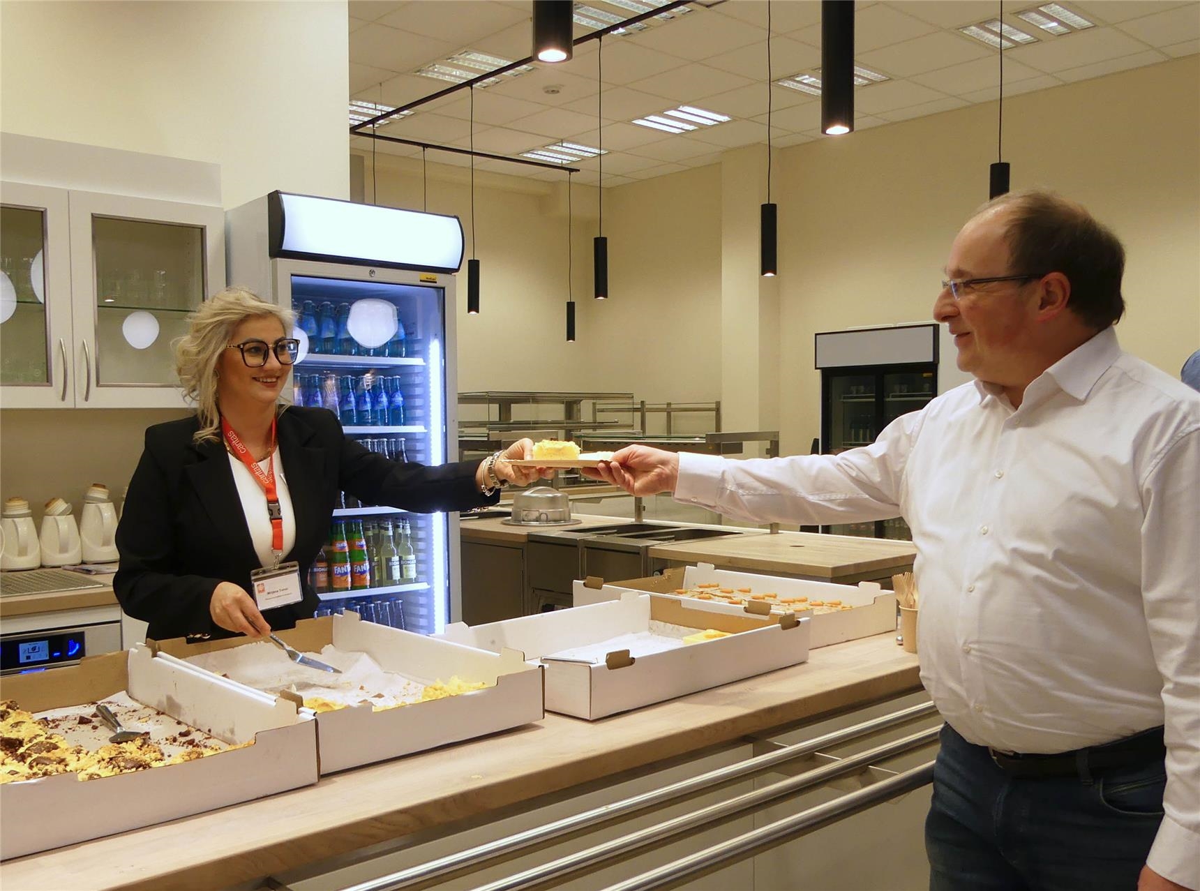 Eine Caritas-Mitarbeitende, die am Kuchenbuffet steht, reicht einem Besucher des Quartierscafés ein Stück Apfelkuchen über die Theke. (Foto: © Sabine Schilha)