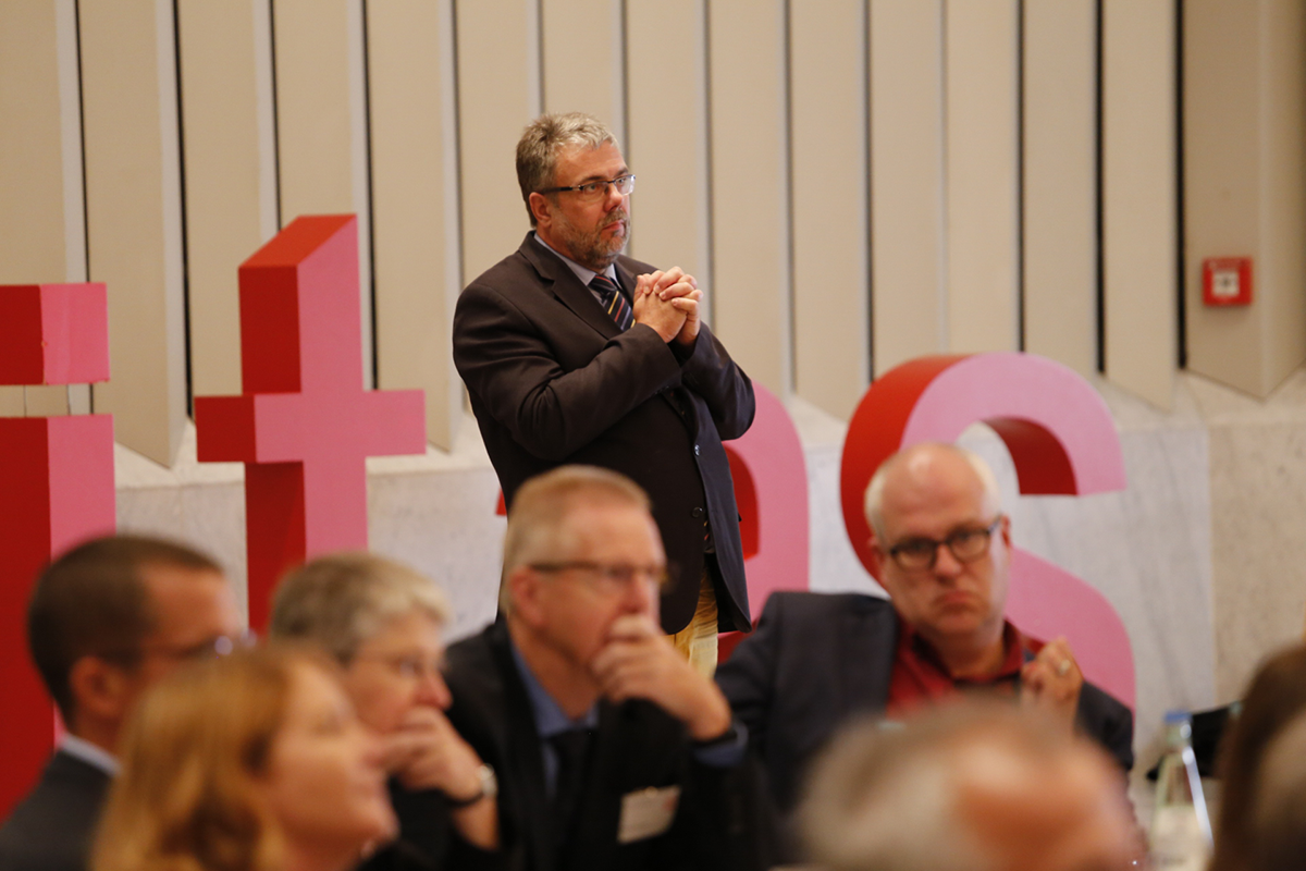 Bruno Schrage steht mit verschränkten Händen hinter dem Publikum des Fachforums 'Von der Kunst, ein christliches Unternehmen zu führen'. Hinter befinden sich große rote Buchstaben.  (Martin Karski)