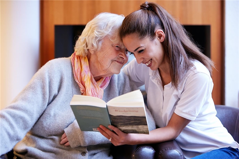 Jüngere Frau und alte Frau blicken gut gelaunt zusammen in ein Buch.