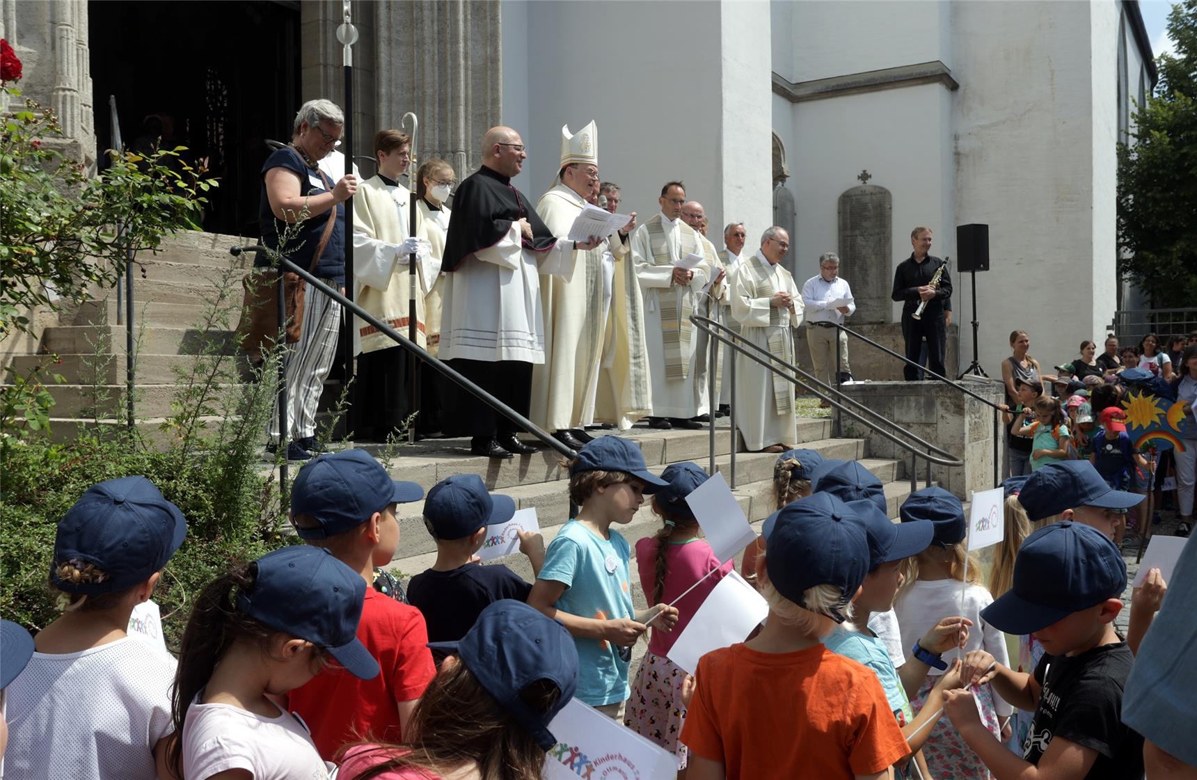 Das Foto zeigt Bischof Dr. Bertram Meier und andere Geistliche, wie sie die Kinder zu ihrer Kinderwallfahrt am Südportal der Ulrichsbasilika begrüßen. 