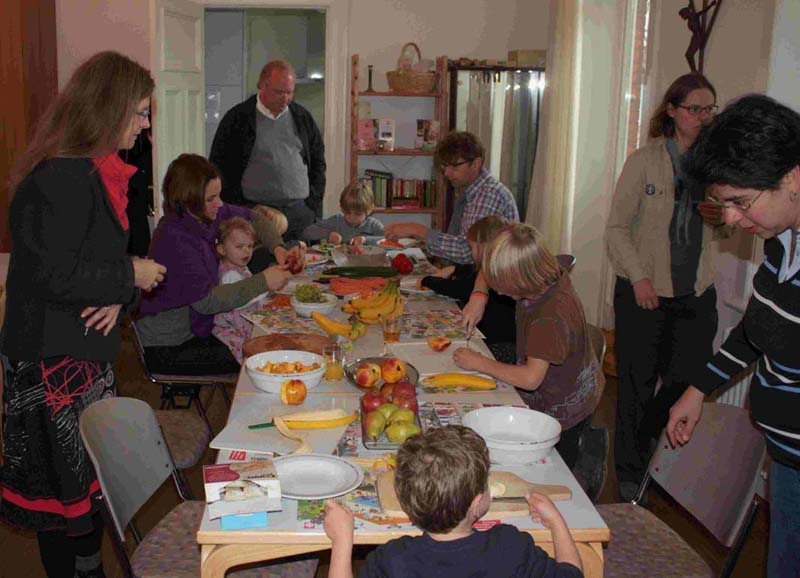Viele Kinder und einige Erwachsene bereiten an einem langen Tisch Obstsalat zu. (Manuela Siegerink-Schröder)