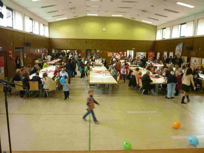 In einer großen hellen Halle sitzen viele Gäste an langen Tischen; Kinder wuseln herum. (Foto: Caritasverband Saar-Hochwald)
