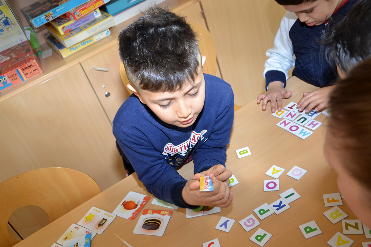Drei Schüler und eine Frau spielen ein, auf einem Holztisch ausgebreitetes Lernspiel in den Räumlichkeiten der Brukterer-Grundschule in Dortmund-Lanstrop. Ein Junge steht dabei im Fokus des Bildes. (Christian Lukas)
