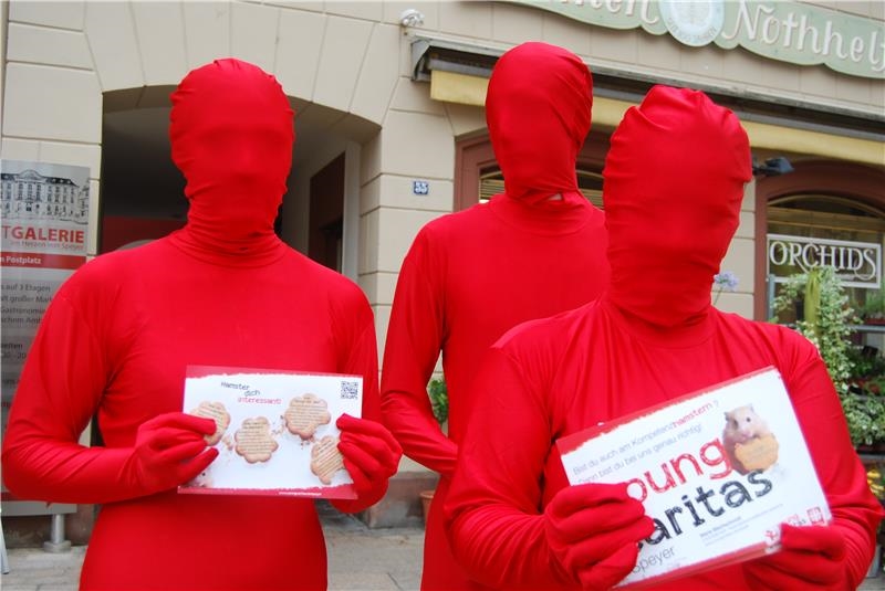 Drei Jugendliche in roten Morphsuits halten Flyer von youngcaritas Speyer in die Kamera. (Caritas Speyer)