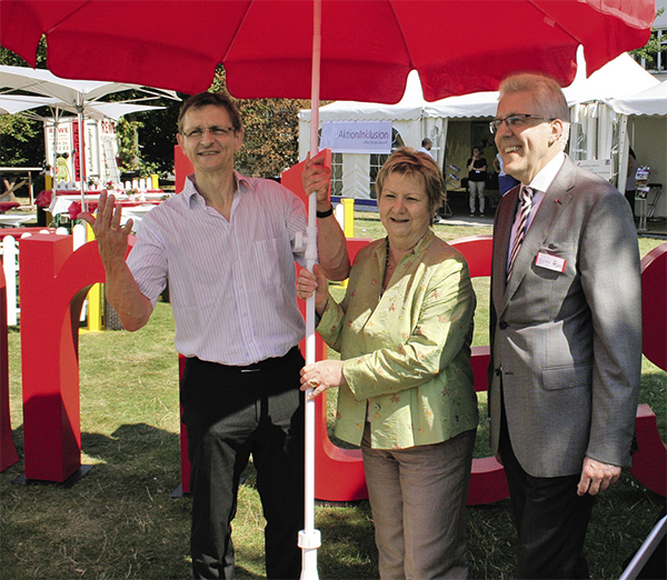 Diözesan-Caritasdirektor Frank Joh. Hensel, Sylvia Löhrmann (Grüne) und Thomas Salmen (OCV Düsseldorf), die auf einer Wiese unter einem roten Caritas-Sonnenschirm stehen (Markus Lahrmann)