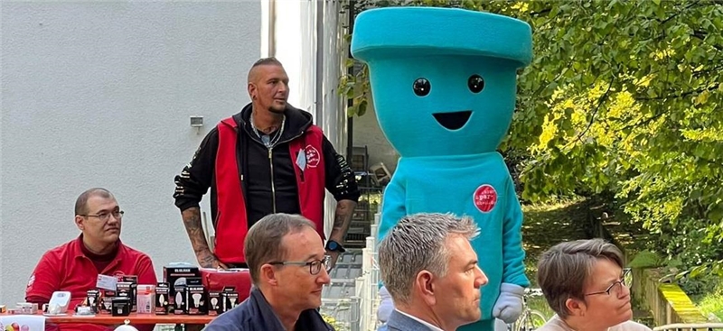 Feier des Stromspar-Checks am 07.10.22 in Waiblingen: Menschen sitzen auf Bänken und schauen in die Richtung der Redner. Im Hintergrund zwei Stromspar-Berater und das Maskottchen, eine Steckdose.