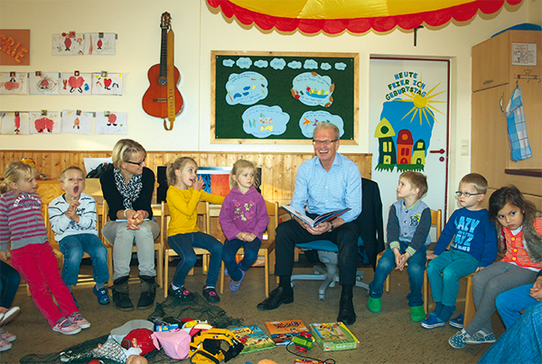 Einige Kinder sowie eine Frau und ein Mann sitzen in einem bunt eingerichten Raum eines Kindergartens im einem Stuhlkreis zusammen. Der Mann hält ein aufgeschlagenes Buch in seinen Händen. (Annika Lacour/Gemeinde Legden)