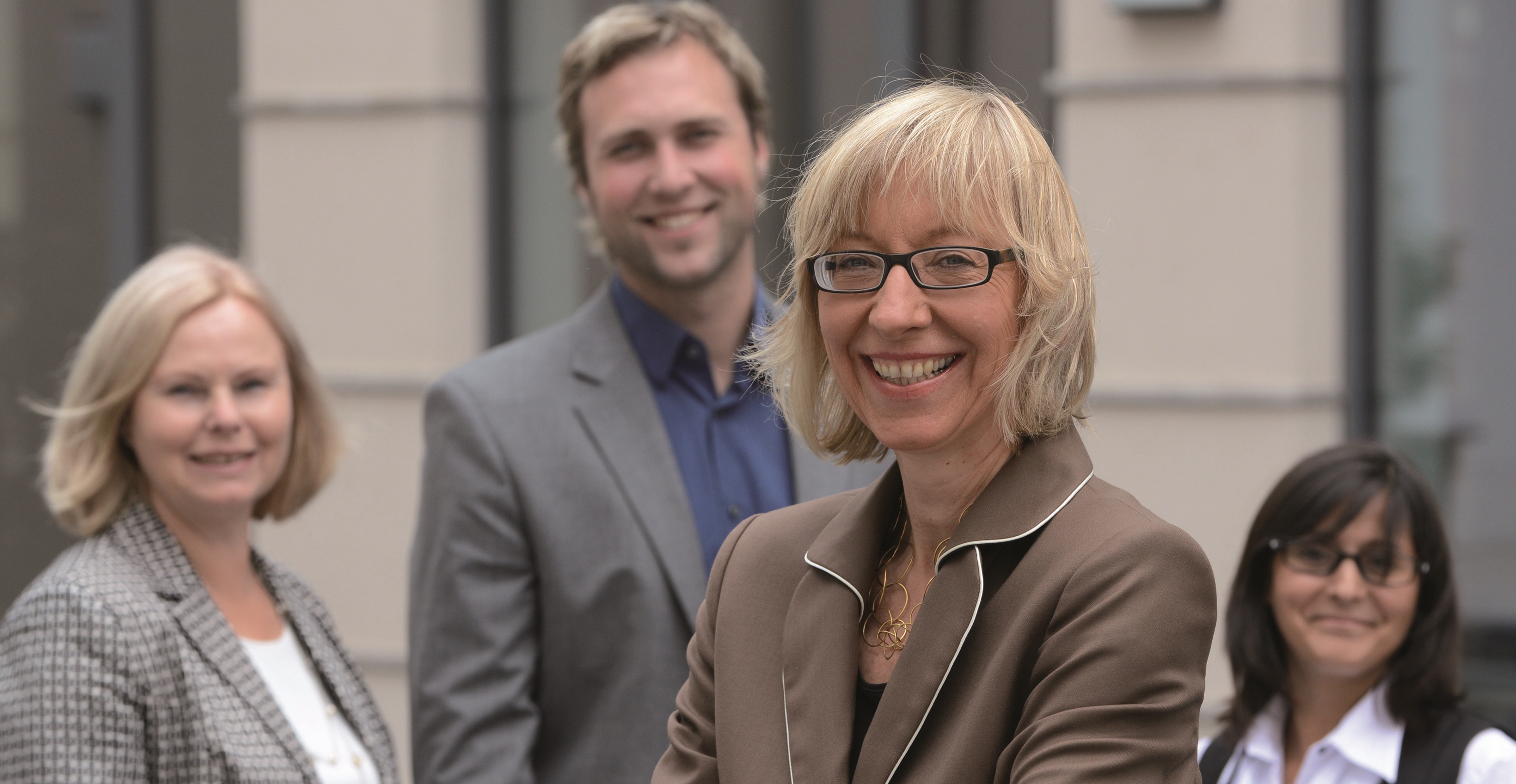 Eine schick gekleidete Frau in einer Führungsposition lächelt in die Kamera, im Hintergrund stehen zwei Kolleginnen und ein Kollege