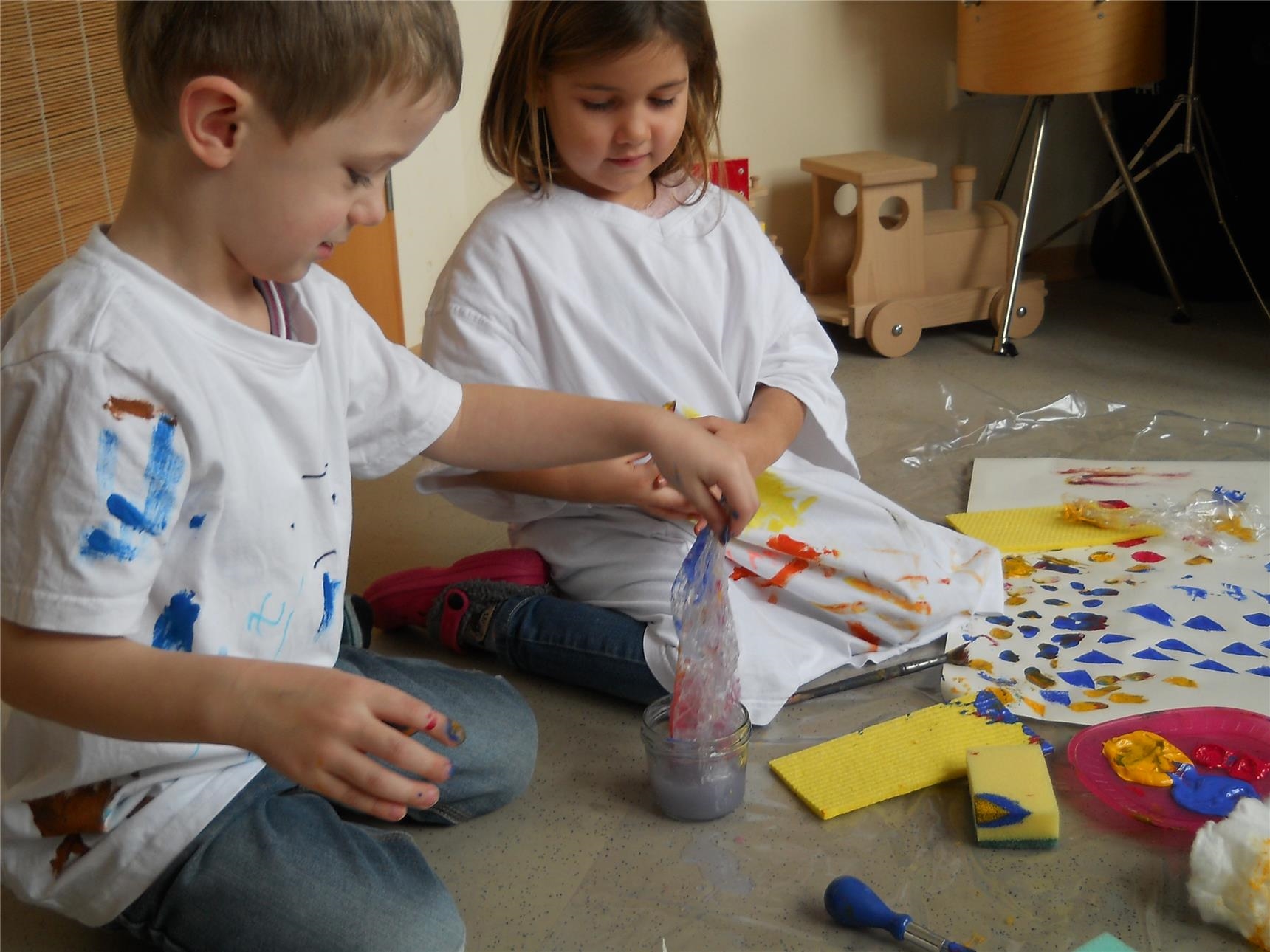 zwei Kinder Knien auf dem Boden, auf dem Papier, Schwämme, Pinsel, Farbe und ein Wasserbehälter verteilt sind (Caritasverband Trier e. V.)