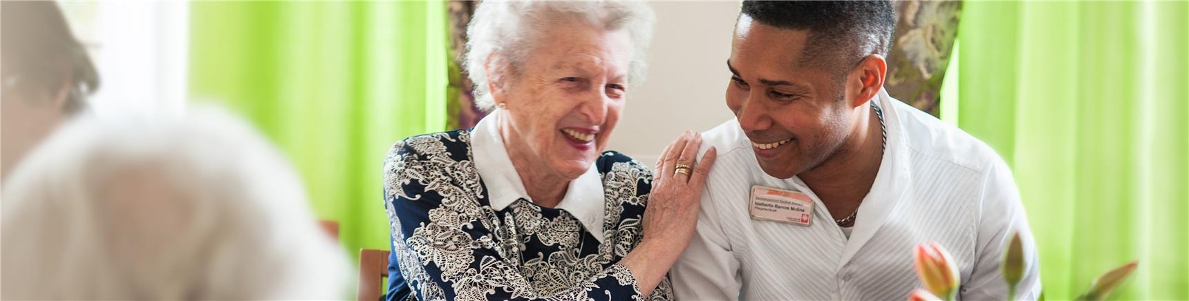 Seniorin und Altenpfleger sitzen nebeneinander am Kaffeetisch und lachen herzlich