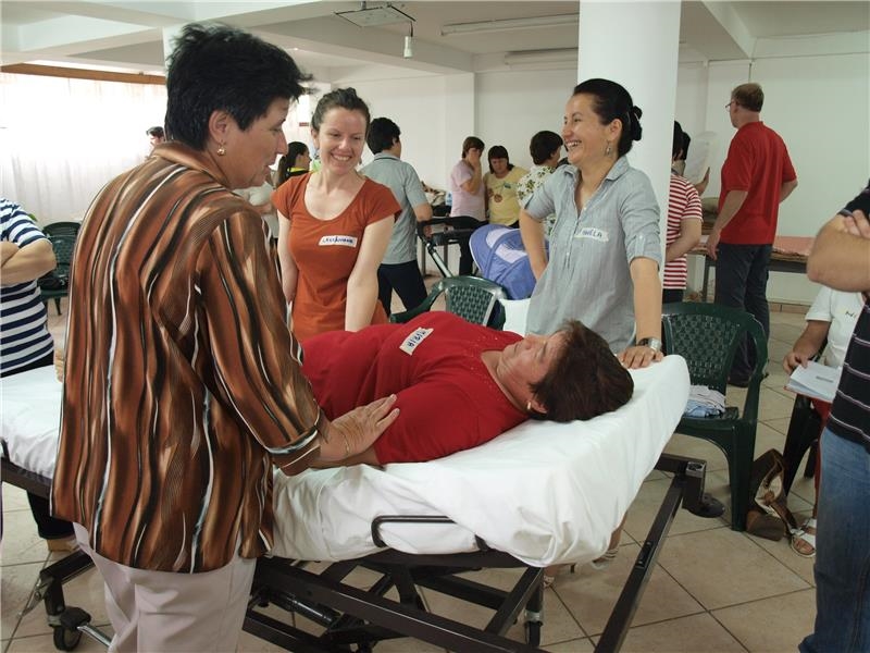 Drei Frauen stehen um ein Krankenbett, auf dem eine Frau liegt. Im Hintergrund sind weitere Personen zu sehen. (Caritas)