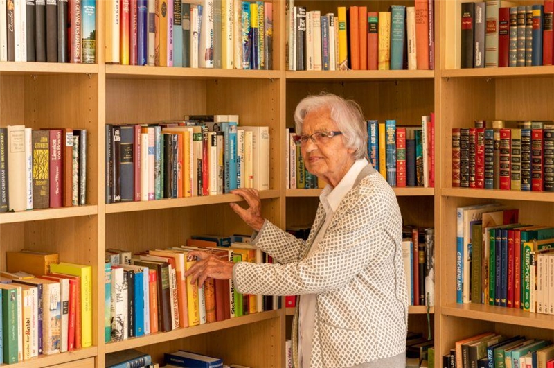 Eine Seniorin steht vor einem sehr großen und bund gefülltem Bücherregal.