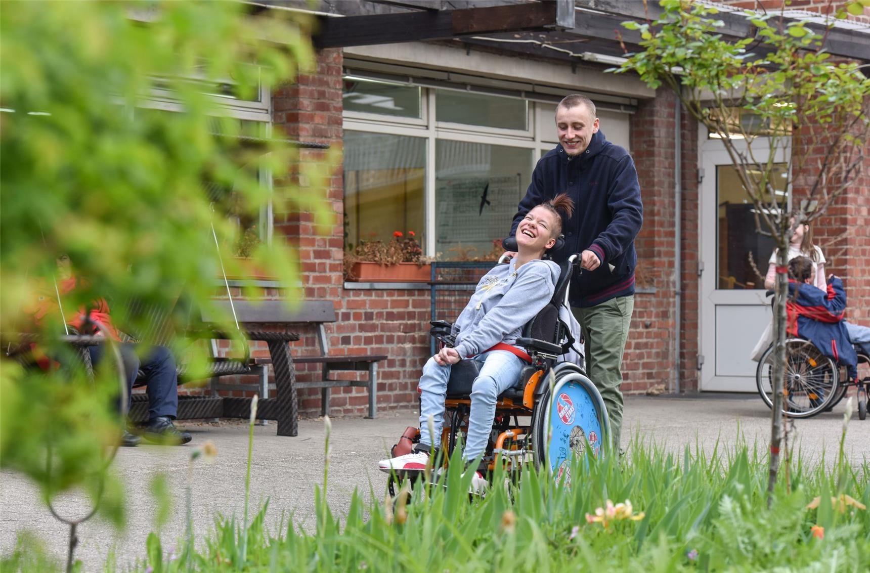 Betreuer schiebt eine junge Frau mit Behinderung auf dem Rollstuhl durch den Garten (Deutscher Caritasverband / Harald Oppitz, KNA)