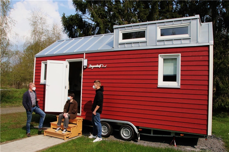 Das Foto zeigt zwei stehende und eine auf den Eingangsstufen eines Tiny House sitzende Person.