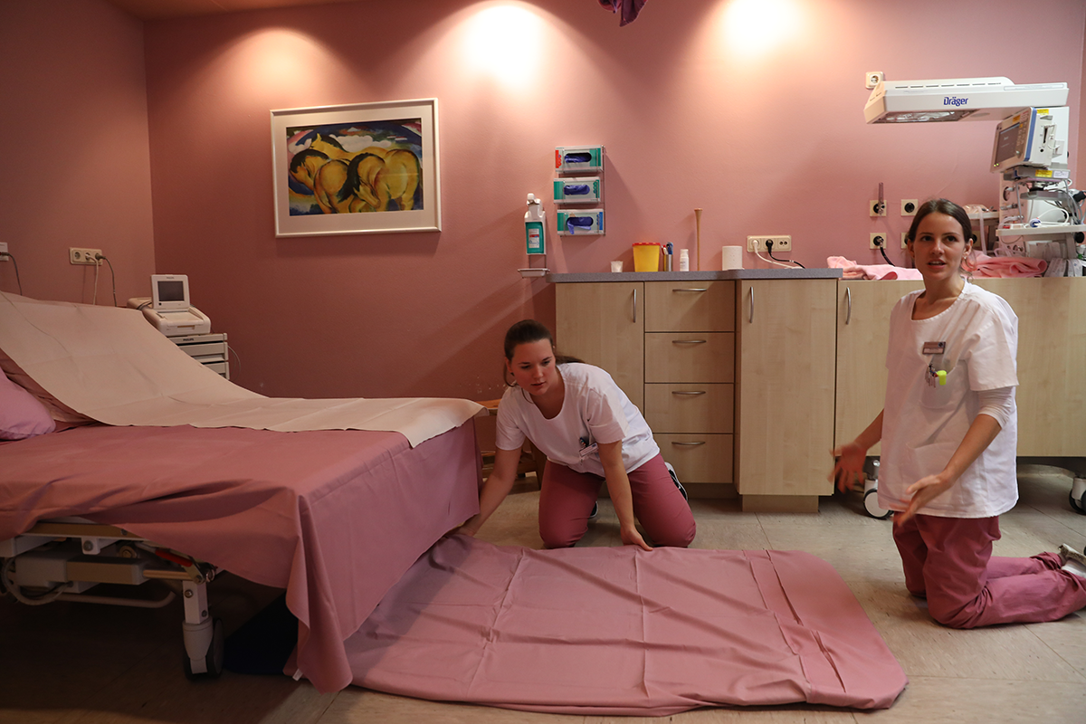 Zwei Hebammen befinden sich in einem Krankenzimmer und knien vor einem Bett auf dem Boden. Eine der Hebammen bereitet eine Decke auf dem Boden aus. (Robert Boecker)