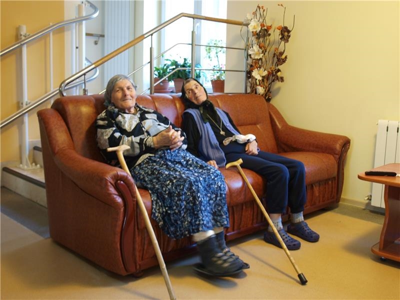 Auf einem Sofa sitzen zwei ältere Frauen, die beide bäuerliche Kleidung und ein Kopftuch tragen (Caritas)