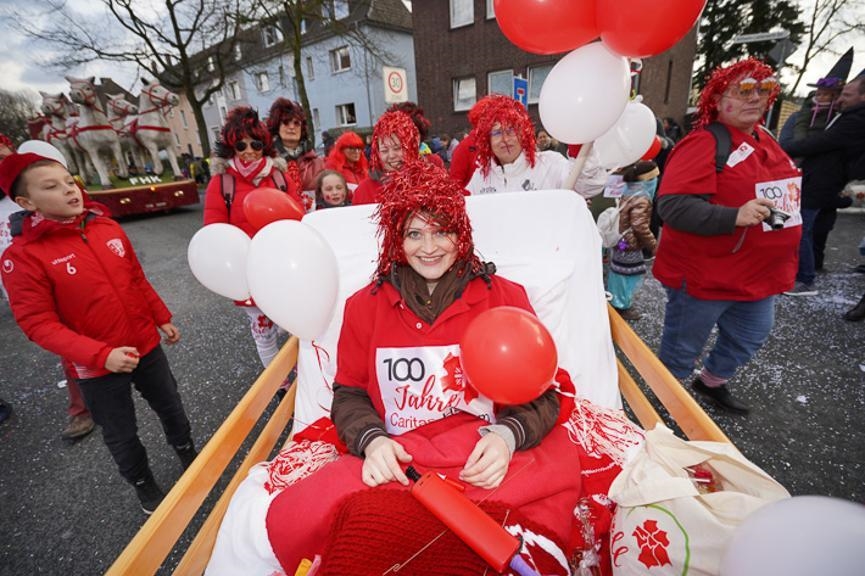 Rot-weiß gekleidete Frau mit roter Perücke liegt im Krankenbett, am Bett sind Luftballons befestigt (WA Szkudlarek)