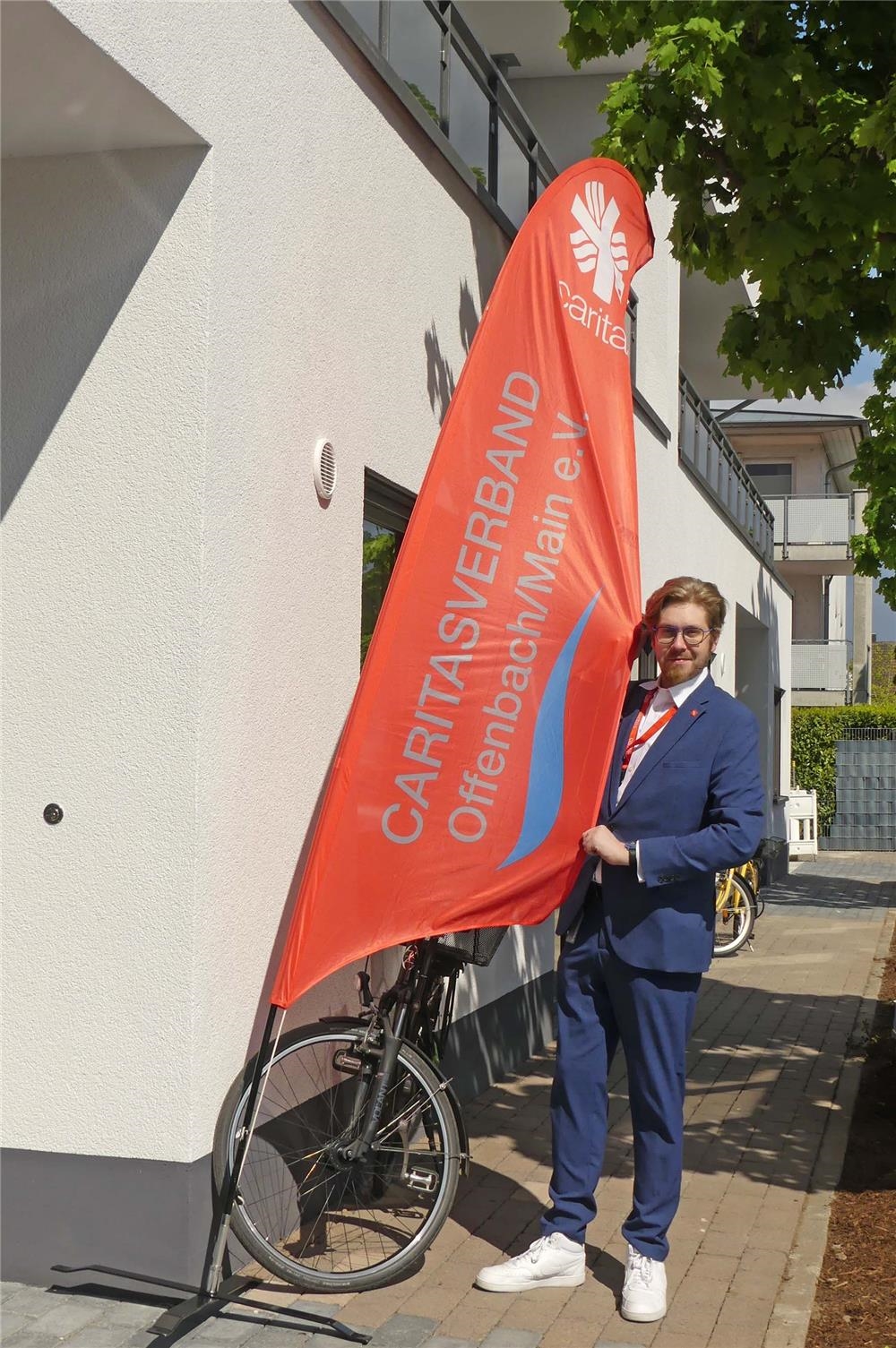 Ein Caritas-Mitarbeiter erwartet Gäste am Eingang von Haus Raphael | Er hält eine rote Caritas-Beachflag fest, die im Wind flattert (Foto: © Caritas Offenbach / Sabine Schilha)