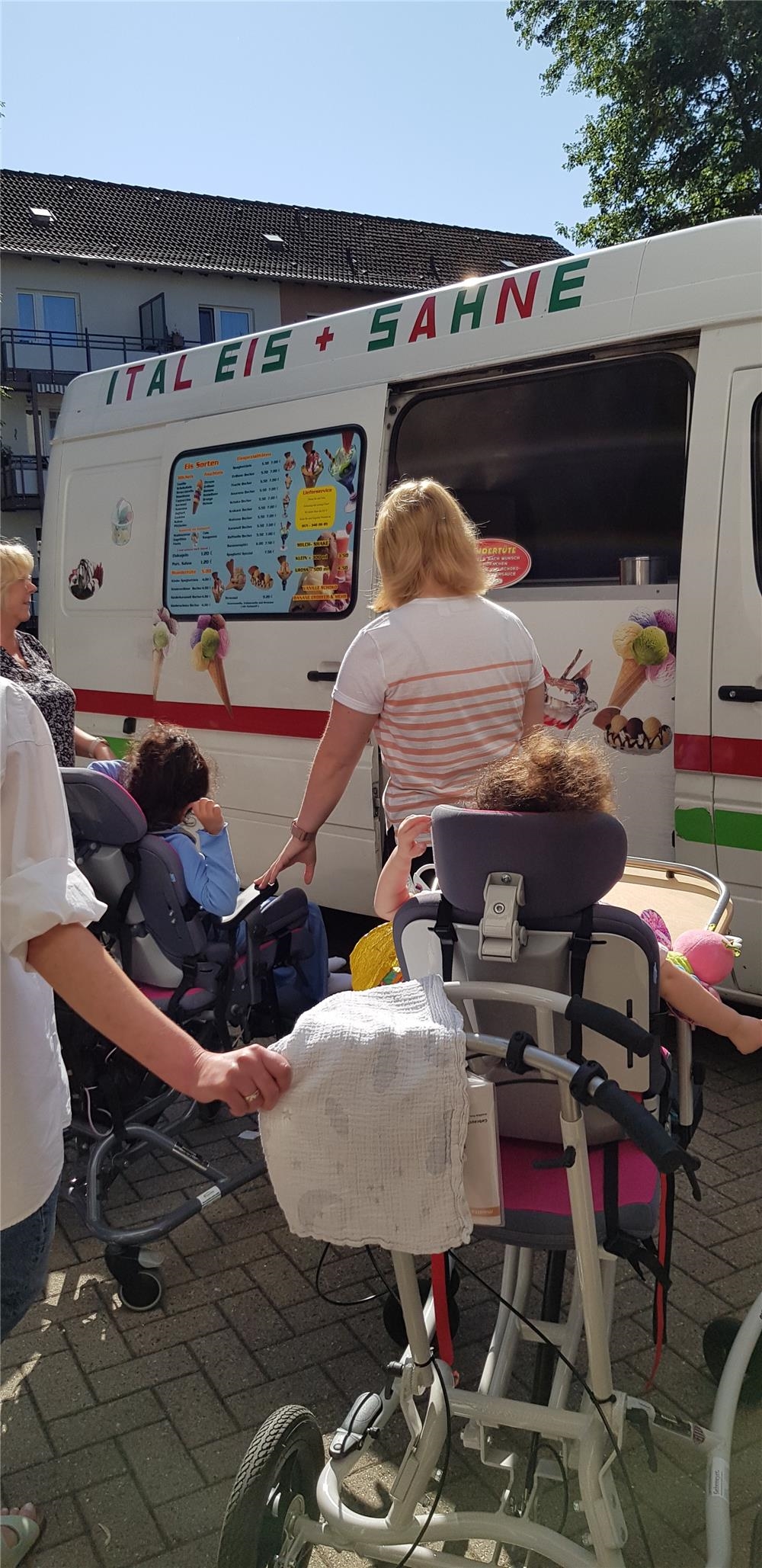 Mitarbeiterinnen und Kinder im Rollstuhl stehen vor einem Eiswagen in der Schlange. 