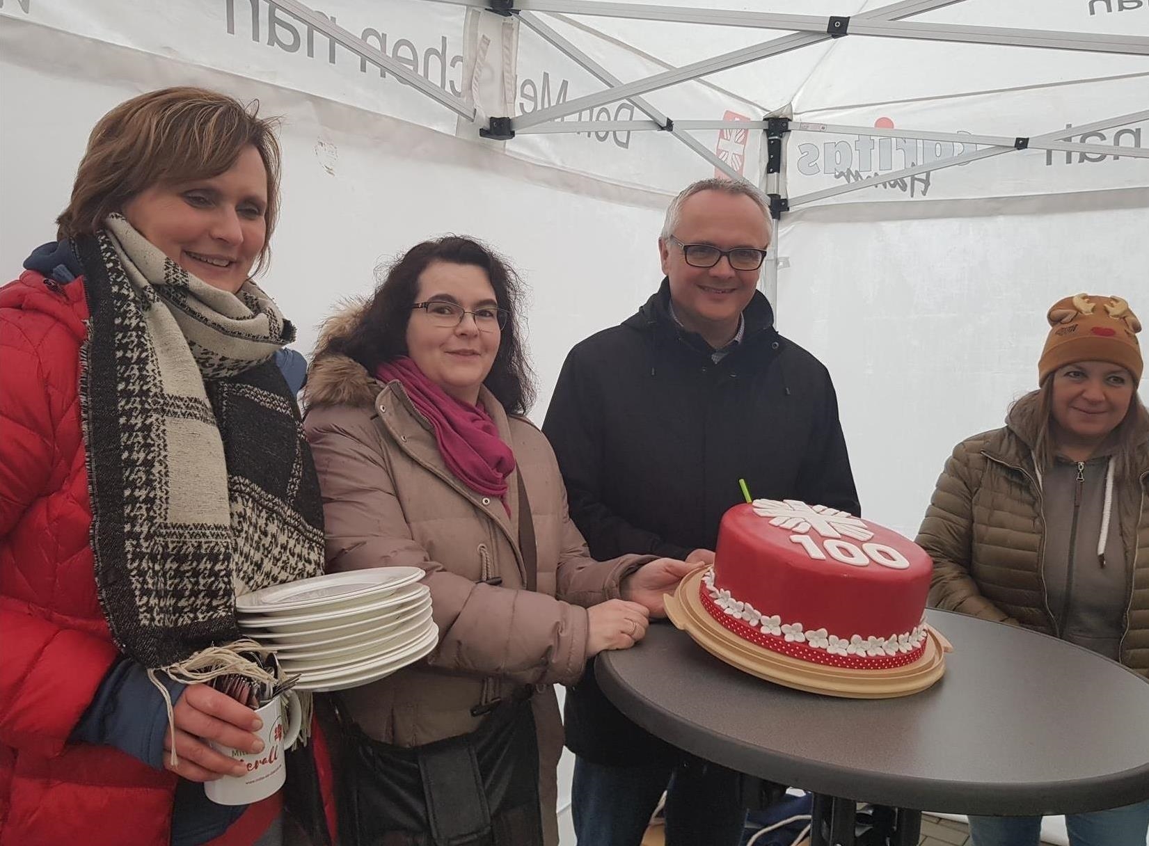 Auf einem Stehtisch steht ein roter Kuchen mit weißem Flammkreuz und der Zahl 100, um den Tisch stehen drei Frauen und ein Mann (Caritas Sozialstation NordWest)