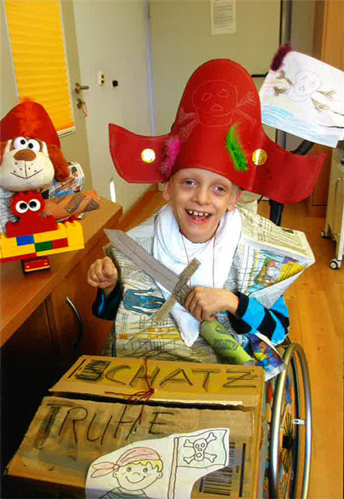 Ein als Pirat verkleideter Junge mit Behinderung sitzt in einem Rollstuhl und hat einen Karton mit der Aufschrift 'Schatztruhe' vor sich stehen. Der Rollstuhl steht in einem Spielzimmer. (Caritas)