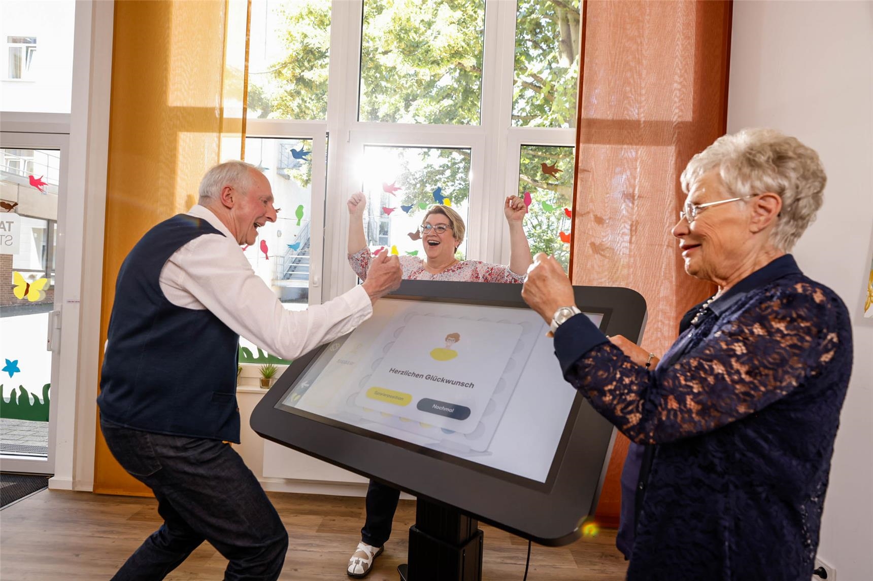 Senioren stehen an einem großen digitalen Tisch, dem care table und jubeln gemeinsam. 