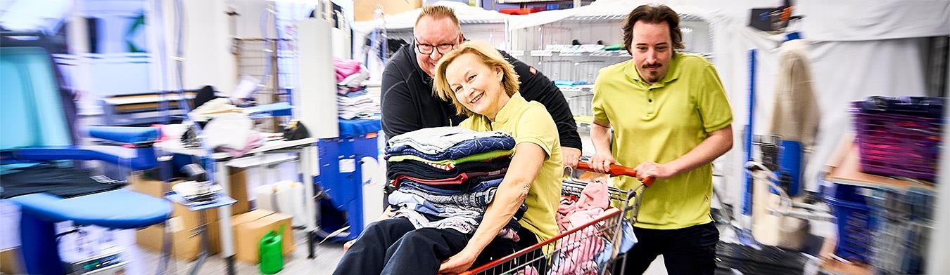 Ein Mitarbeiter der Wäscherei schiebt eine Frau in einem Einkaufswagen durch die Wäscherei.