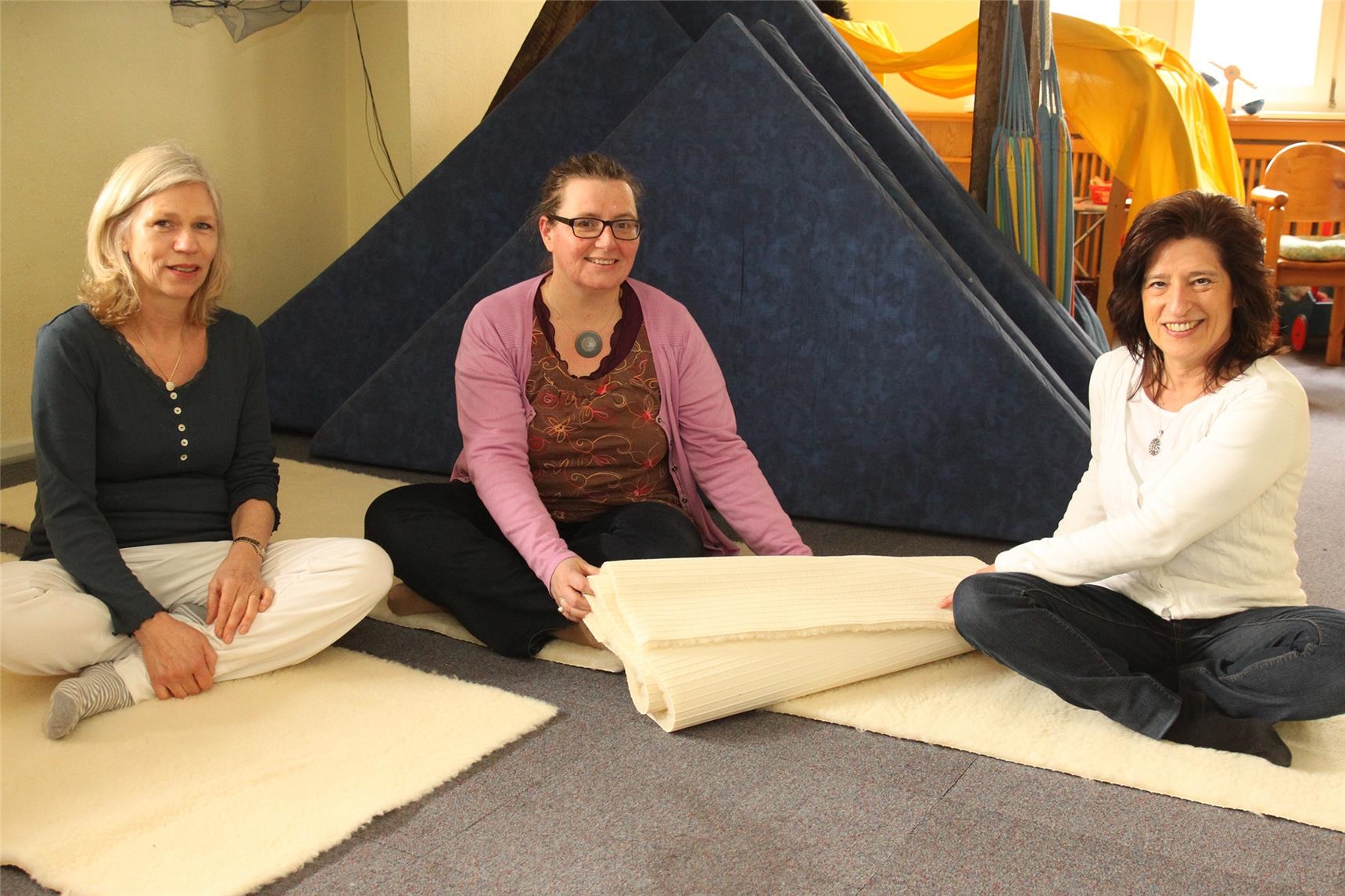 Eine Frau übergibt eine Yoga-Matte; insgesamt sitzen drei Freunde auf drei Matten im Schneidersitz 