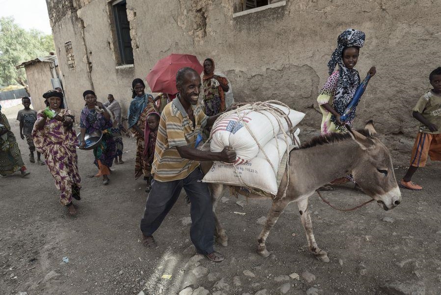 Ein Esel trägt Säcke, daneben laufen lachend Menschen. (Christoph Gödan, Caritas international)