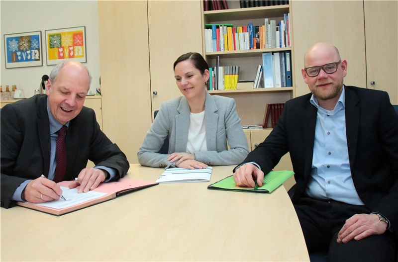 Das Foto zeigt zwei Männer und eine Frau an einem Tisch, der Mann links unterschreibt einen Vertrag.